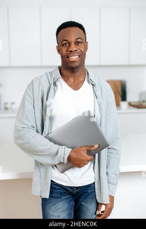 Porträt eines afroamerikanischen, gutaussehenden Mannes, der seinen Laptop in der Hand hält, eines schwarzen Studenten oder Freiberuflers, in stilvoller Freizeitkleidung, der zu Hause in der Küche steht, die Kamera anschaut, lächelt freundlich Stockfoto