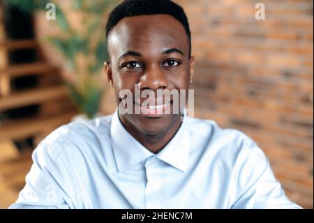 Nahaufnahme eines afroamerikanischen, positiv erfolgreichen Geschäftsmanns, Managers, Freiberuflers, Büromitarbeiters, der ein Hemd trägt, Sitzen in einem modernen Büro, lächeln freundlich an der Kamera Stockfoto