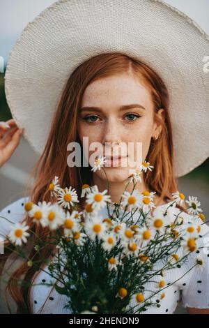 Nahaufnahme eines Rothaarigen mit Hut und einem Strauß Daisen an sonnigen Sommertagen. Frau im Kamillenfeld. Natürliche Schönheit. Glückliche Menschen Stockfoto