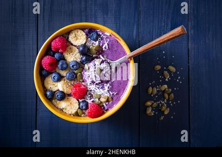 Gesunde Heidelbeer-Smoothie-Schale mit Banane, Himbeere, Kürbiskern, Hanfsamen und Kokosnuss in einer gelben Schale auf einer blauen Holzoberfläche Stockfoto