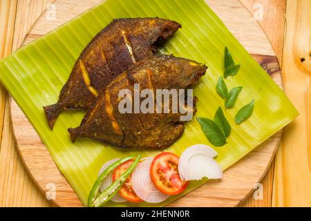 Pomfret-Fischbraten, garniert mit Tomatenscheiben, Zwiebelscheiben und Zitronenscheiben, die in einem Bananenblatt auf Holzboden angeordnet sind. Stockfoto