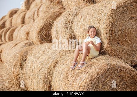 Porträt eines lächelnden jungen Mädchens mit Sundress, das auf einem hohen Heuhaufen sitzt und sich auf einen anderen und viele andere Heuhaufen im Hintergrund lehnt. Haben Stockfoto