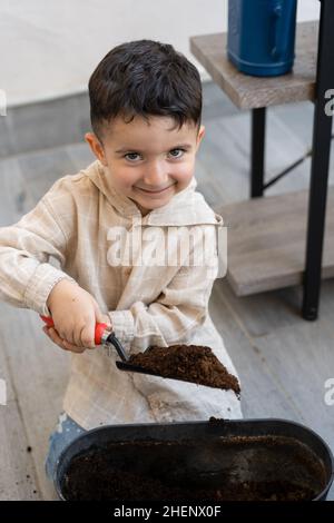 Lustiger Junge pflanzt zu Hause ein kleines Kind mit Schaufel setzt Erde in den Blumentopf und verpflanzt Pflanze A in einen neuen Topf. Innenpflege, Biophilie-Design und Liebe für Zimmerpflanzen. Stockfoto