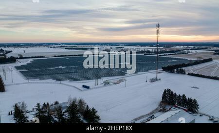 Am späten Nachmittag werden in einer verschneiten ländlichen Gemeinde eine Solarfarm und ein Zellenturm am Horizont gesehen. Stockfoto