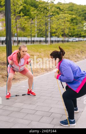 In voller Länge vertikale Porträt von übergewichtigen jungen Frau tut Kniebeugen Übungen mit Fitness-Band für die Gewichtsabnahme mit Personal Trainer im Freien. Stockfoto