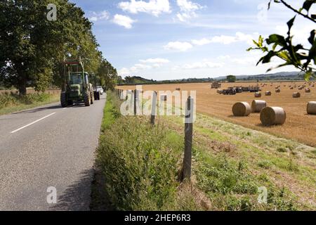 Erntezeit in Somerset, England. Ein Traktor fährt auf einer Straße neben einem Feld voller Heuballen Stockfoto