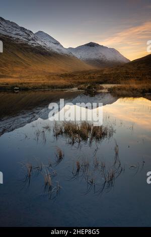 Glencoe Schottland, Lochan na Fola, stehend in Hochlandsmooren und tiefem Gras, schneebedeckte Berge links und rechts und eine Spiegelreflexion auf dem Wasser. Stockfoto