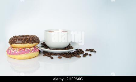 Aromatischer Kaffee-Latte mit Blatt auf der Oberseite in weißer Tasse auf dem Teller, bedeckt mit braunen Samen neben den Donuts. Snacks aus Schokolade und Himbeere Stockfoto