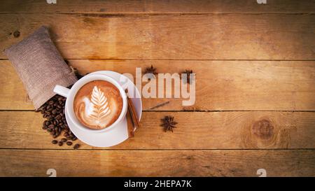 Blick auf die Kaffeetasse von oben auf einem alten Holztisch. Blattmuster auf Cafe Latte. Anissterne, Kaffeebohnen im Beutel und Zimt zur Dekoration. Schön, organisch, Stockfoto