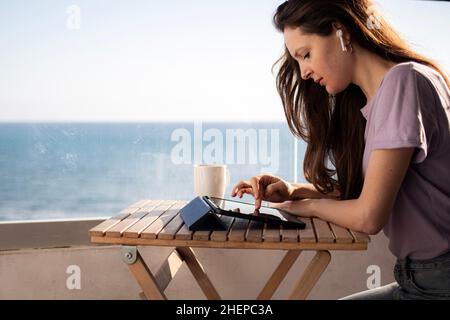 Eine junge Frau mit Kopfhörern arbeitet am Tablet auf dem Balkon in der Nähe des Meeres Stockfoto