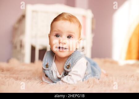 Das 5 Monate alte Neugeborene liegt auf dem Bauch auf dem Bett. Baby Mädchen lächelt und spielt im Zimmer. Denim-Sundress. Weiche Bettüberwürfe. Stockfoto