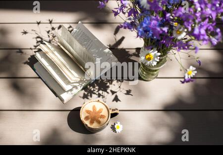 Guten Morgen, romantische Atmosphäre, Komfort, Ruhe, Entspannung zu Hause. Eine Tasse Kaffee mit Milch, eine Vase mit wilden Fliederblumen, ein offenes Buch o Stockfoto