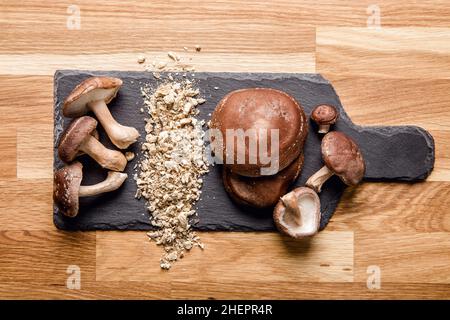 Flache Ansicht von trockenem Pulver aus Shiitake-Pilzen, Lentinula edodes. Lebensmittelzutat auf Schneidebrett aus schwarzem Stein mit frischen Shiitake-Pilzen. Stockfoto