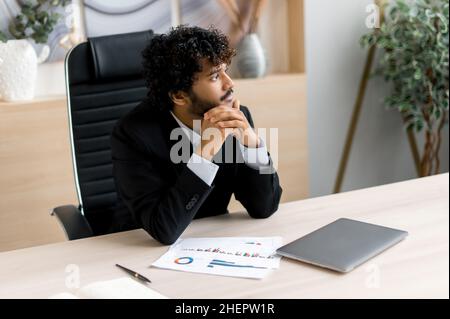 Erfolgreiche attraktive nachdenkliche indische Geschäftsführerin, Top-Manager oder CEO in formeller Kleidung mit, sitzen an einem Tisch im Büro, ruhen von der Arbeit, wegschauen, denken über zukünftige Geschäftspläne Stockfoto