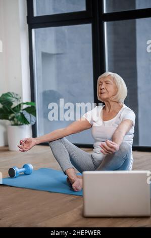 Grauhaarige ältere Frau, die auf dem Boden sitzt und einen Online-Yoga-Kurs macht Stockfoto