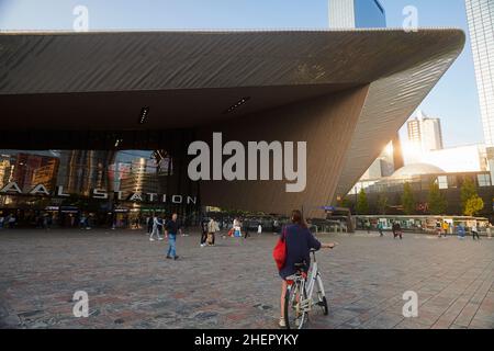 Junge Frau mit Fahrrad wartet vor dem Eingang des modernen Rotterdamer Hauptbahnhofs bei strahlendem Sonnenschein Stockfoto