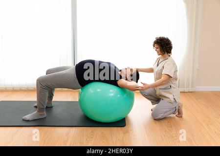 Nahaufnahme einer Schwangeren mit Gesichtsmaske, die auf dem Boden sitzt und mit einem Ball Pilates-Übungen mit einem Physiotherapeuten macht Stockfoto