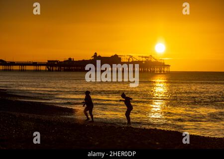 Brighton, West Sussex, 12/01/2022, Einheimische Schwimmer, die von einem frühen Morgenbad am Brighton Beach mit dem Palace Pier im Hintergrund während des Sonnenaufgangs in Brighton, West Sussex, zurückkehren Stockfoto