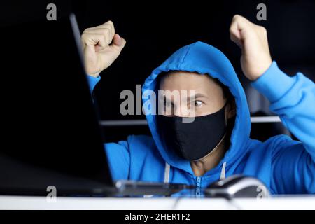 Freude und Überraschung, Mann in schwarzer Maske und blauem Hoodie, der mit den Händen nach oben am Laptop sitzt. Gewinnen Sie die Lotterie oder Computerspiel Stockfoto