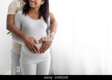 Lächelnder junger afroamerikanischer Mann umarmt den Bauch einer Schwangeren, macht das Herz mit den Händen im Innenraum unerkennbar Stockfoto