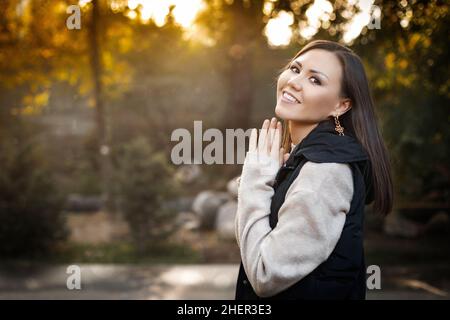 Lächelnde junge Frau auf der rechten Seite des Fotos steht im Herbst im Freien in einem warmen Pullover und einer ärmellosen Jacke. Hochwertige Fotos Stockfoto