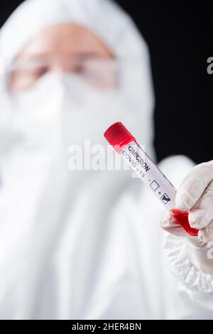 Nahaufnahme des positiven Covid-19-omicron-Variantentests in der Hand eines verschwommenen Arztes in einem Hazmat-Anzug isoliert auf schwarzem, stockem Bild Stockfoto