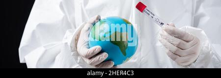 Beschnittene Ansicht des Arztes in einem Hazmat-Anzug mit Globus und Covid-19-Reagenzglas isoliert auf schwarzem Banner, Stockbild Stockfoto