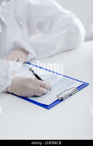 Teilansicht des verschwommenen Arztes in einem Gefahrgutanzug, der in einer medizinischen Karte auf der Zwischenablage im Labor geschrieben wurde, Stockbild Stockfoto