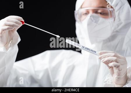 Verschwommener Wissenschaftler im Hazmat-Anzug, der Covid-19 omicron-Variant-Abstrichtest isoliert auf schwarzem, stockem Bild hält Stockfoto