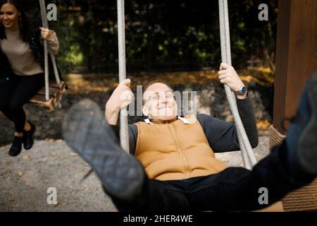 Ein junger, fröhlicher Mann schwingt auf einer Schaukel mit hochgedrehten Beinen und seinem Geliebten im Hintergrund in einem Waldpark. Hochwertige Fotos Stockfoto