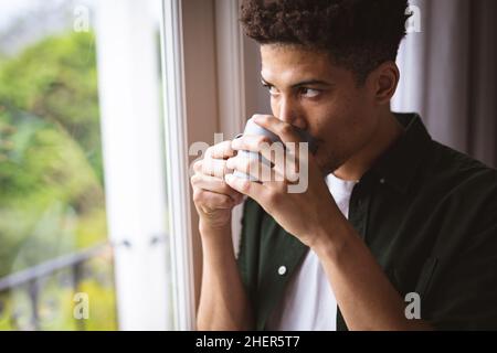 Nachdenklicher junger Birazialmann, der Kaffee trinkt, während er zu Hause durch das Fenster blickt Stockfoto