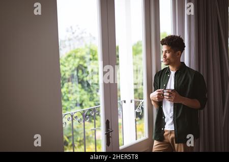 Nachdenklicher junger Birazialmann, der Kaffeebecher hält, während er zu Hause durch das Fenster blickt Stockfoto