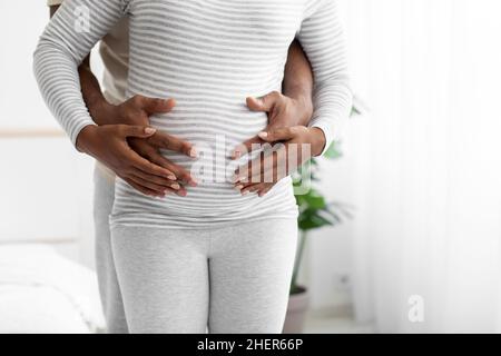 Glücklicher Tausendjähriger afroamerikanischer Mann umarmt den Bauch einer Schwangeren im Schlafzimmer Stockfoto