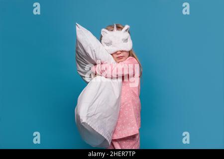 Süßes europäisches blondes Mädchen in rosa Pyjamas und mit einer Einhorn-Maske auf ihren Augen steht auf blauem Hintergrund. Das Kind hält ein großes weißes Kissen, Stockfoto