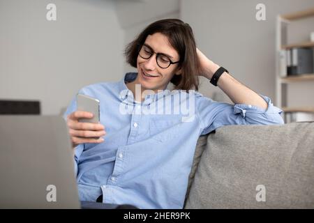 Fröhlicher junger Mann, der Video-Chats mit Geliebten genießt, auf den Smartphone-Bildschirm schaut und sich ausruht, während er am Laptop arbeitet Stockfoto