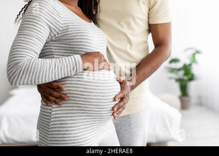 Der ausgeschnittene, tausendjährige afroamerikanische Mann umarmt den Bauch einer Schwangeren im Schlafraum, leerer Raum Stockfoto