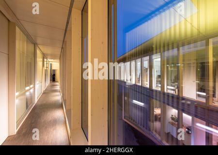 Blick vom Gang eines Bürogebäudes aus Holzkonstruktion durch das Fenster zur Glasfassade bei Nacht Stockfoto