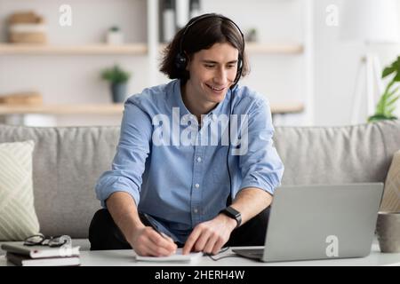 Online-Bildungskonzept. Fröhlicher junger Mann im Headset, der zu Hause mit einem Laptop studiert und sich beim Webinar Notizen macht Stockfoto