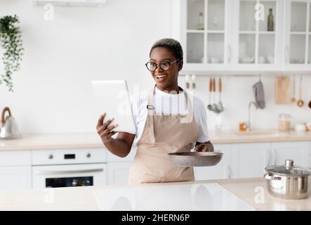 Überrascht glückliche junge afroamerikanische Frau in Schürze und Brille schaut auf Tablet und bereitet das Abendessen zu Stockfoto