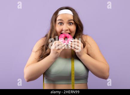 Porträt von lächelnd hungrig lustig tausendjährigen europäischen Frau plus Größe in Sportuniform mit Maßband essen Donut Stockfoto