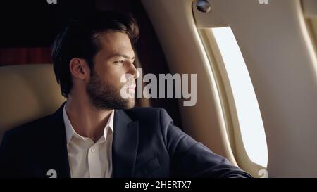 Bärtiger Geschäftsmann, der im Privatflugzeug auf das Fenster blickt Stockfoto