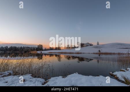 Farm auf einem Hügel am See im Winter, während der orange Himmel des Sonnenuntergangs Stockfoto