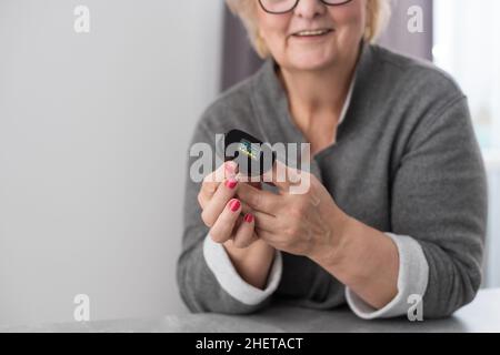 Lächelnde ältere Frau, die zu Hause das Pulsoximeter verwendet, die Sauerstoffsättigung selbst misst, die Pulsfrequenz überprüft und während der Messung Gesundheitsindikatoren kontrolliert Stockfoto