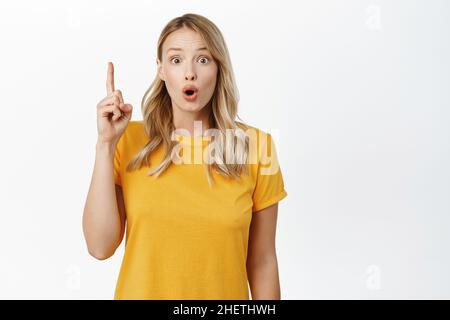 Staunendes blondes Mädchen, das Werbung zeigt, mit dem Finger nach oben zeigt, etwas oben zeigt, Logo oder Banner voraus, weißer Hintergrund Stockfoto