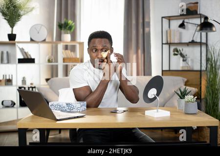 Porträt eines hübschen afrikanischen Mannes, der zu Hause am Tisch sitzt und goldene Anti-Falten-Flecken unter seinen Augen aufführt. Telearbeit vom Heimbüro, Hygiene zu Hause Stockfoto
