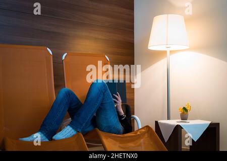 Das Mädchen liest ein Buch, das in einer Liege mit Lampe vor einer Holzwand liegt Stockfoto