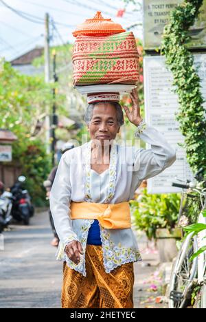 BALI, INDONESIEN - 14. APRIL 2018. Lächelnde balinesische Frau in traditioneller Kleidung, die einen Korb trägt. Redaktionelle Tagesaufnahme Stockfoto