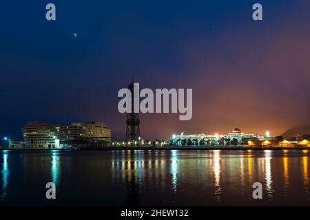 Küstenstadtbild bei Nacht mit bunten Strahlen von reflektierten Lichtern, die über das Wasser tanzen, und einem Leuchten am Himmel durch die Beleuchtung der Stadt Stockfoto