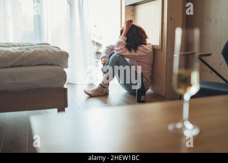 Traurige Frau, die mit einer Flasche Alkohol auf dem Boden im Schlafzimmer saß. Unfokussiertes Weinglas auf dem Vordergrundtisch. Psychische Gesundheit und Alkoholismus pro Stockfoto