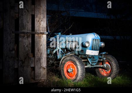 Blauer Oldtimer-Traktor, der nachts neben einer Holzhütte mit rot lackierten Reifen steht Stockfoto